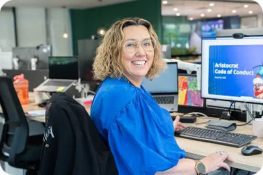Mujer sonriendo desde su escritorio en un entorno de oficina, con un ordenador que muestra el "Código de Conducta Aristócrata